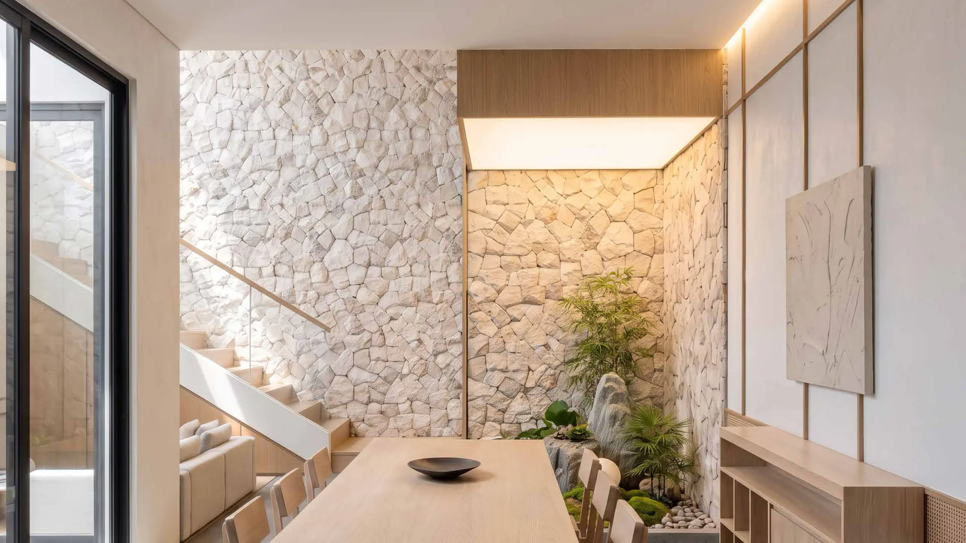 A double-height courtyard clad in rough white stone with a floating staircase and dining table.